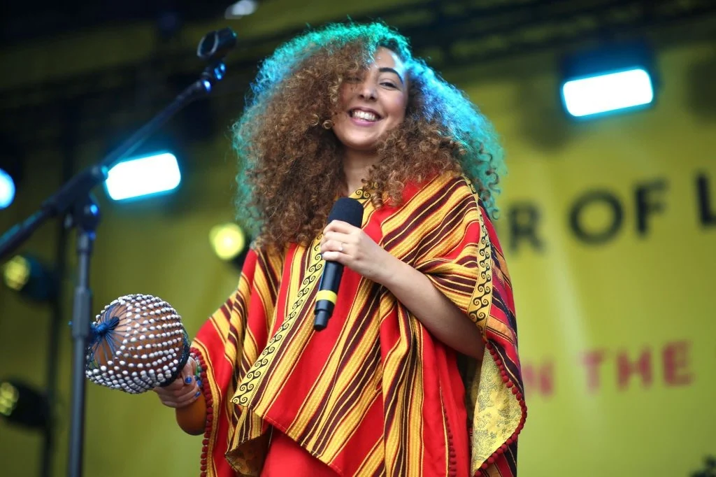 Music for All ambassador Kenza Blanca singing into a microphone on an outdoor stage, holding a frame drum