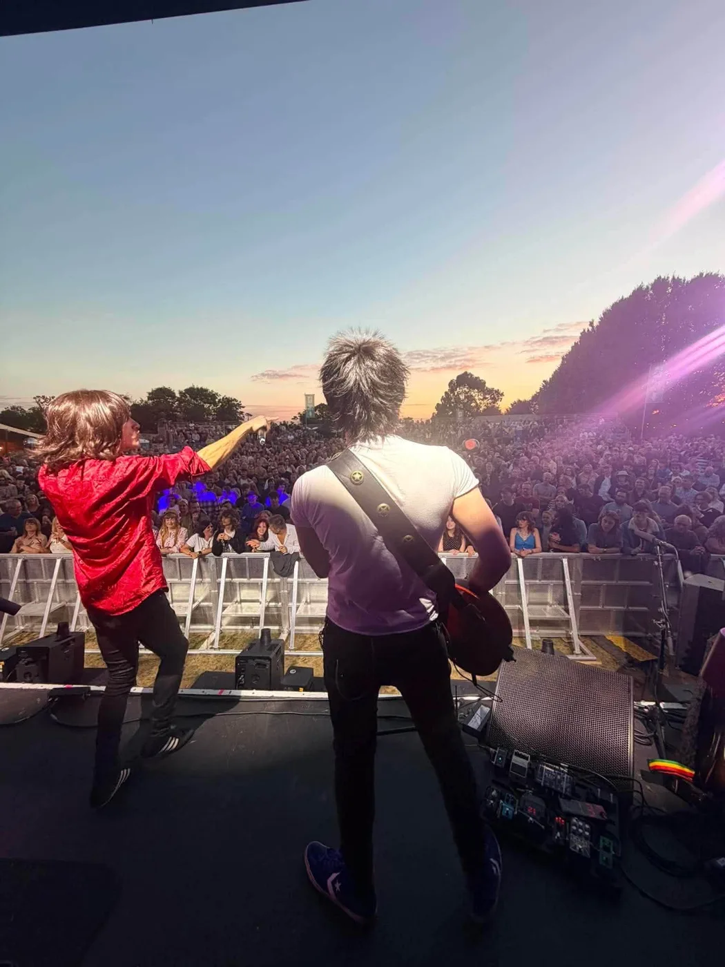 Two members of The Rolling Clones, seen from behind on stage, one in a red shirt pointing at a large outdoor crowd under a sunset sky, the other playing guitar