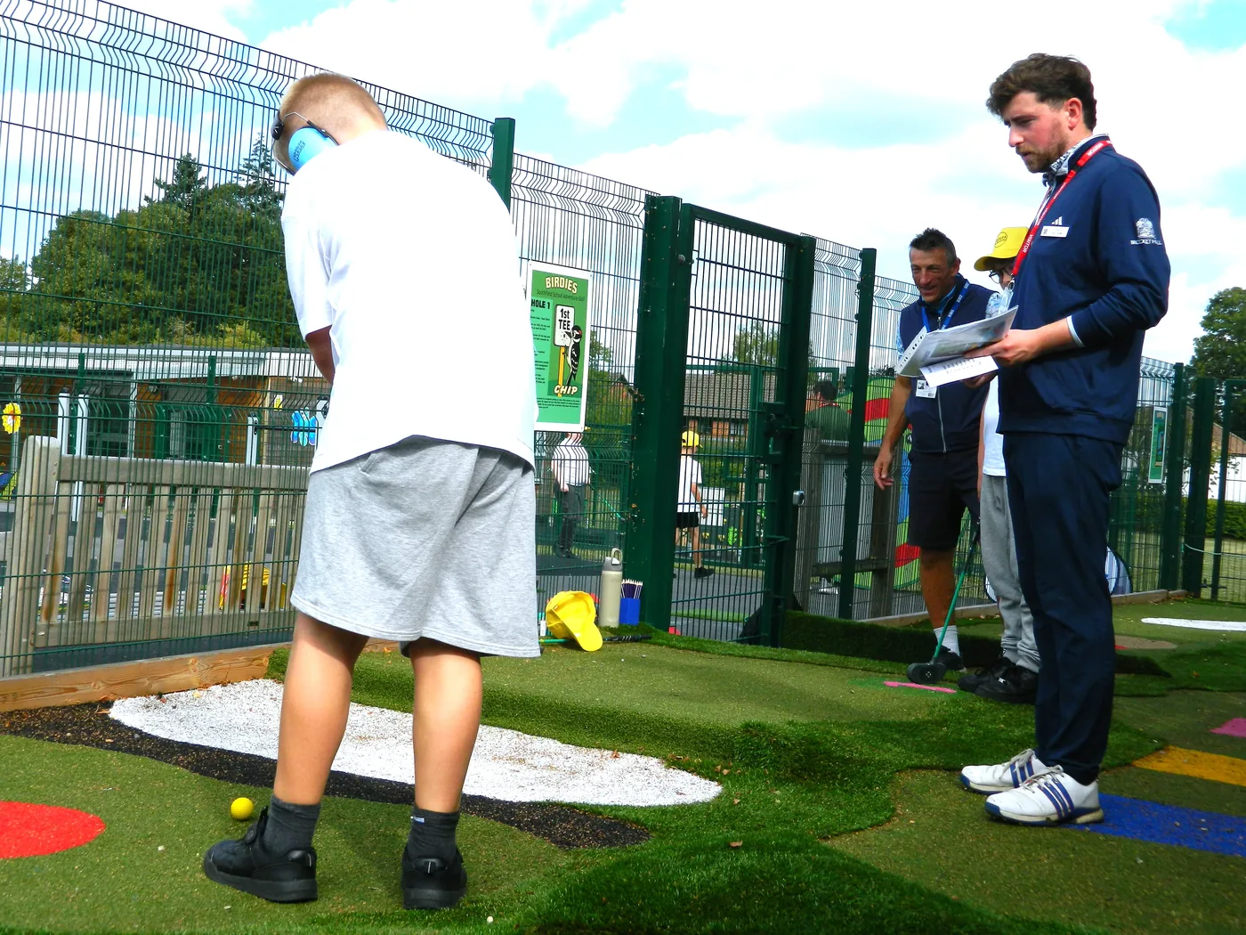 A pupil standing over a ball at a hole while a man in a navy tracksuit holds a sheet of paper and another adult watches