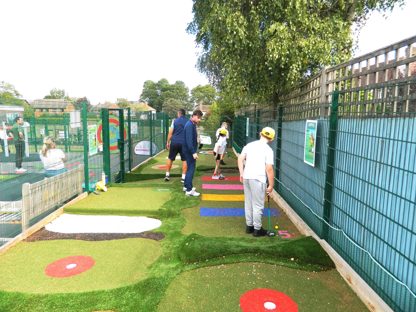 A long narrow adventure golf course running beside a green school fence, with pupils and adults playing along it